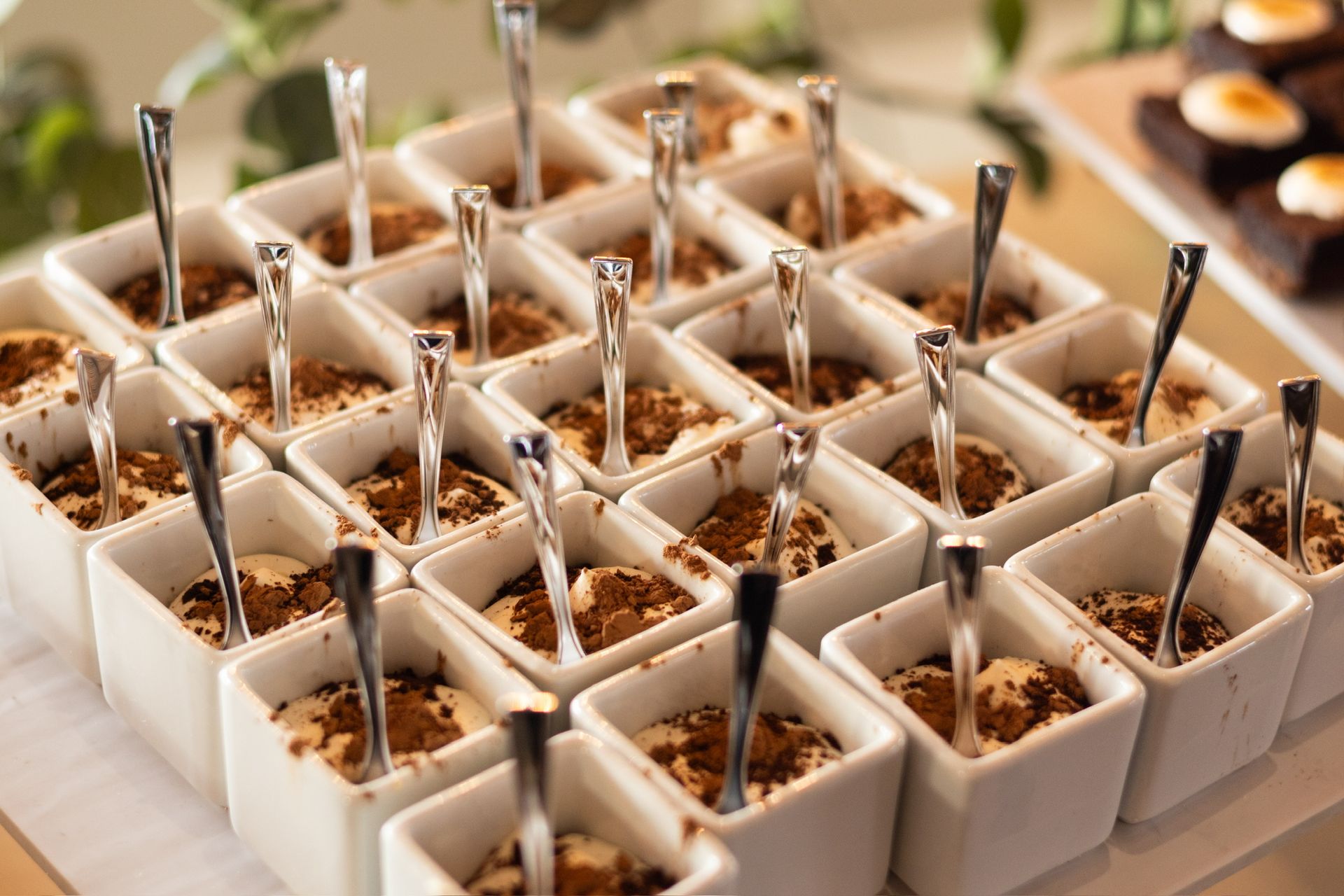 Rows of individual square white dessert dishes, each with creamy filling, cocoa powder, and a small spoon.