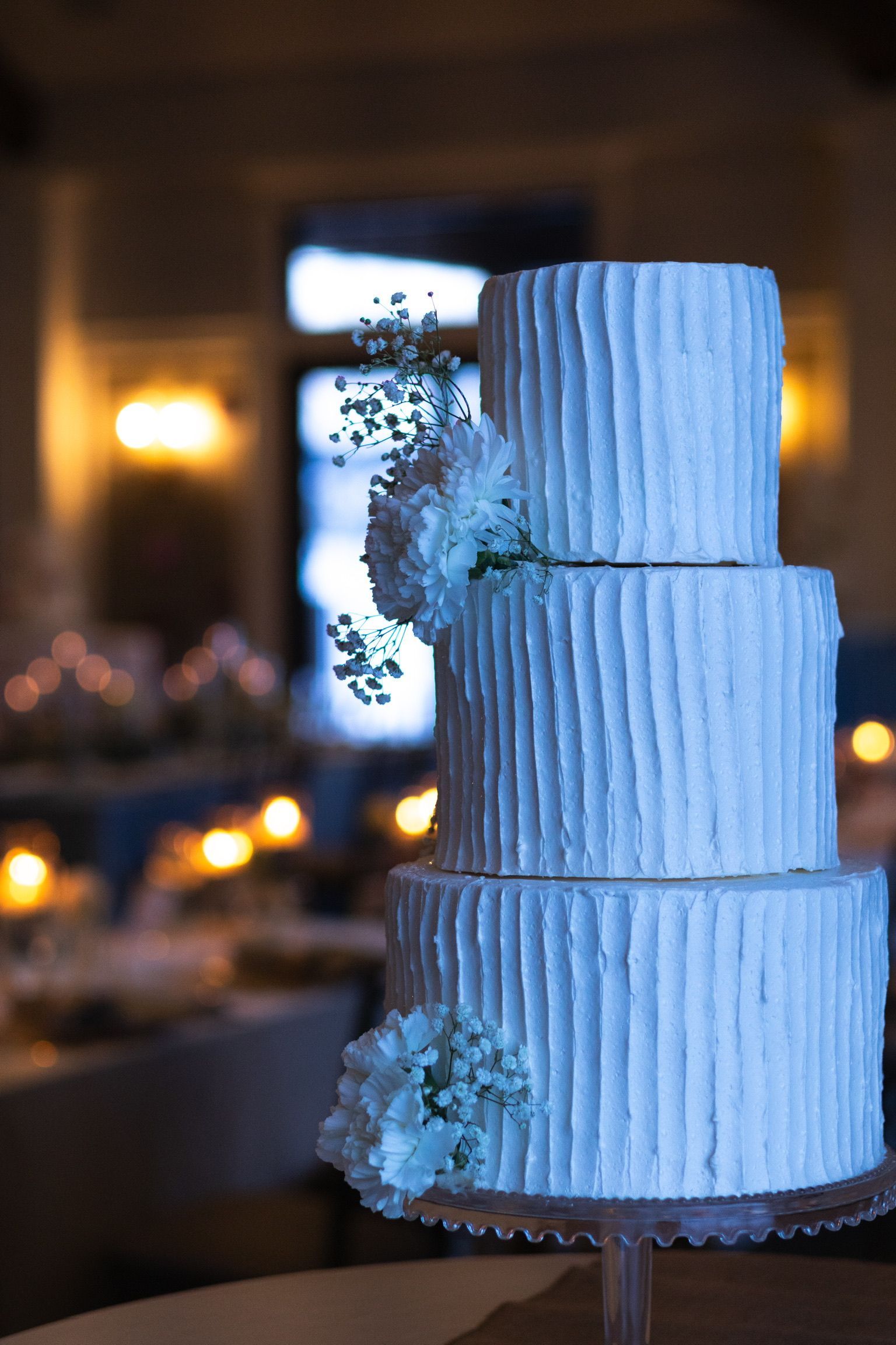 Three-tiered white wedding cake with textured frosting, decorated with small flowers. Lit with soft background lights.