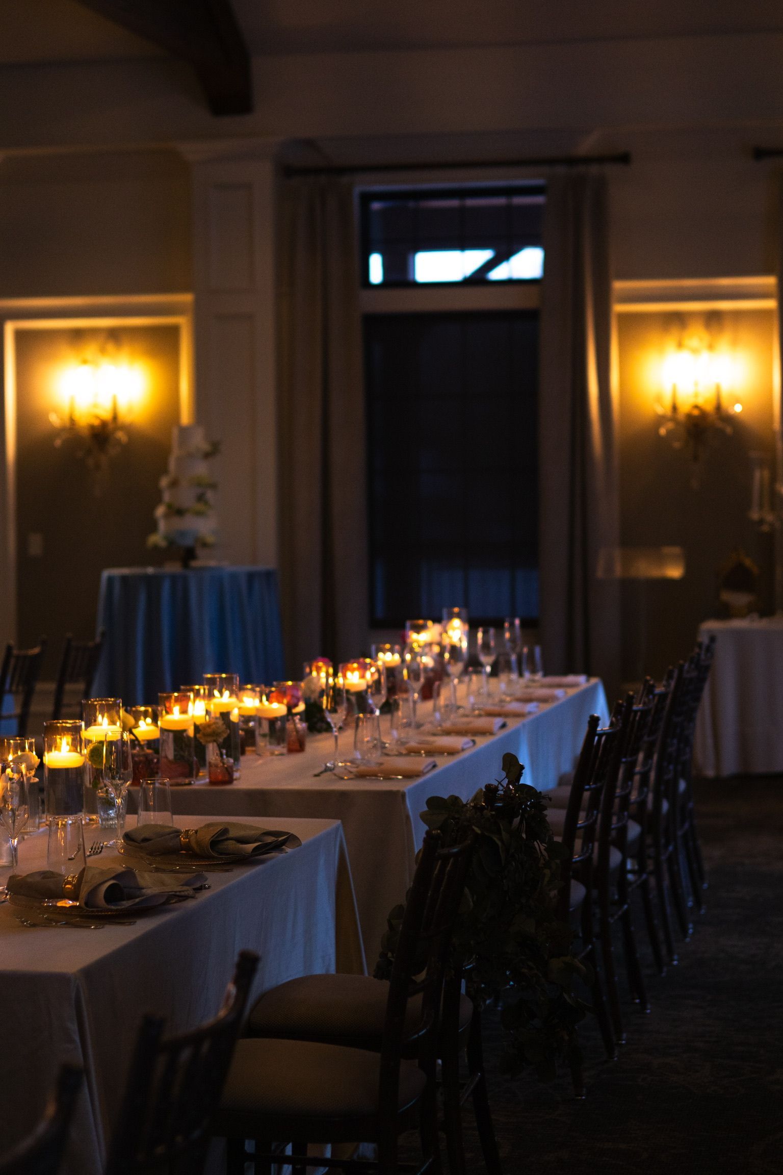 Long, candlelit dining table set for a formal event, with dark chairs and cake visible in the background.