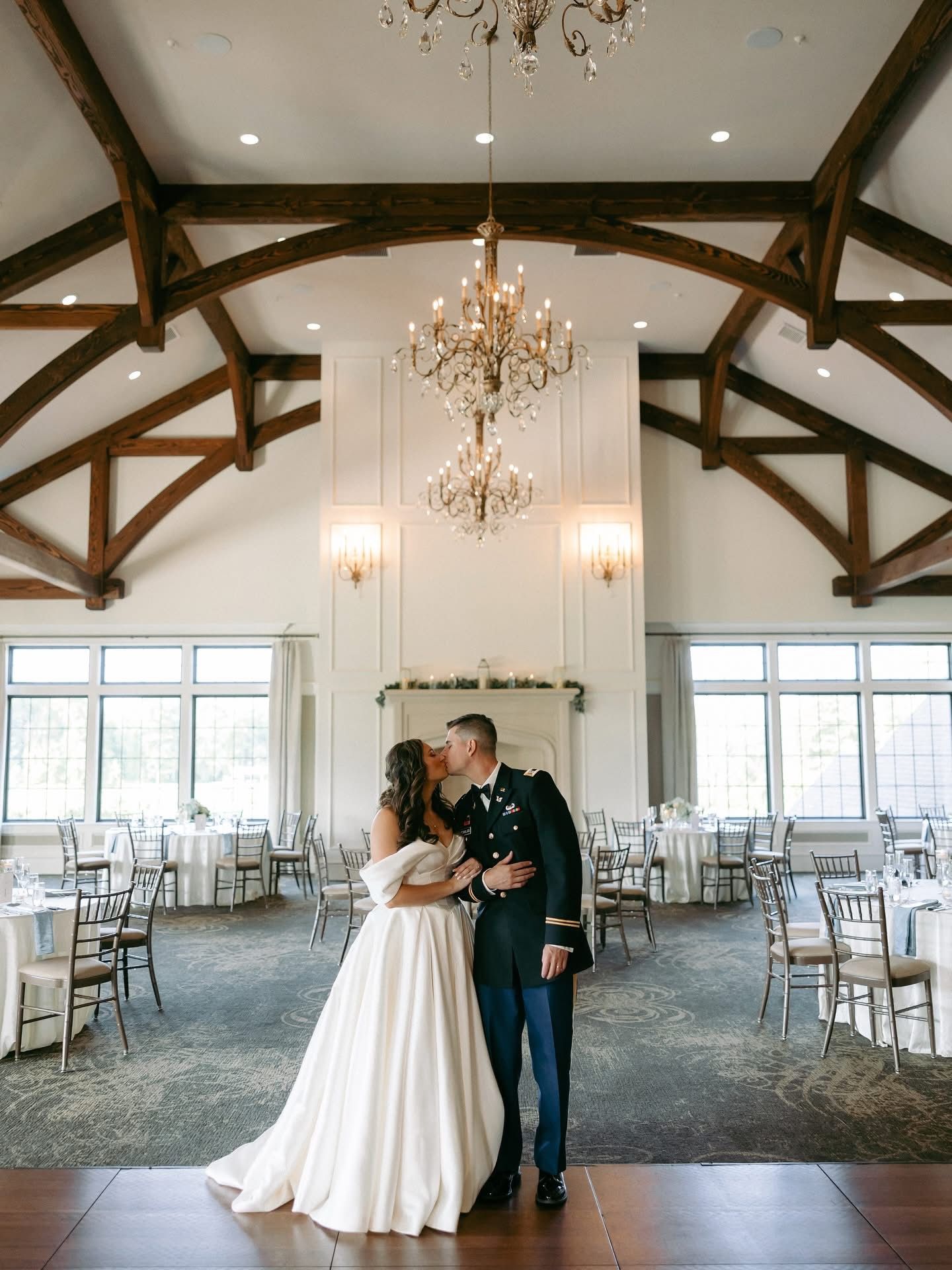 Bride and groom kissing, formal attire, in a wedding reception hall with chandeliers and tables.