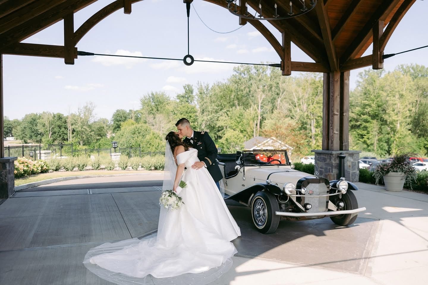 Bride and groom kissing by a vintage car, under a wooden structure; outdoor wedding setting.