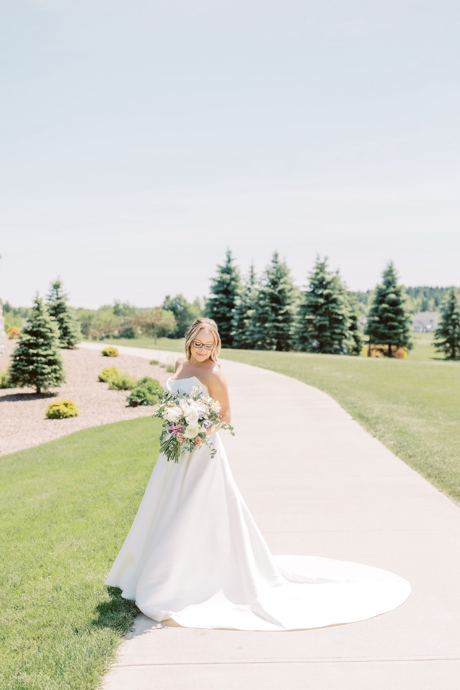 Bride in white gown holding bouquet, smiles on sunny path lined with trees.