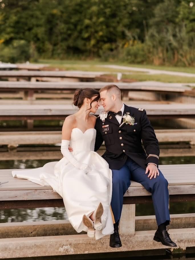 Bride and groom in formal attire sit close, heads touching, on wooden dock. They smile, with water and foliage background.