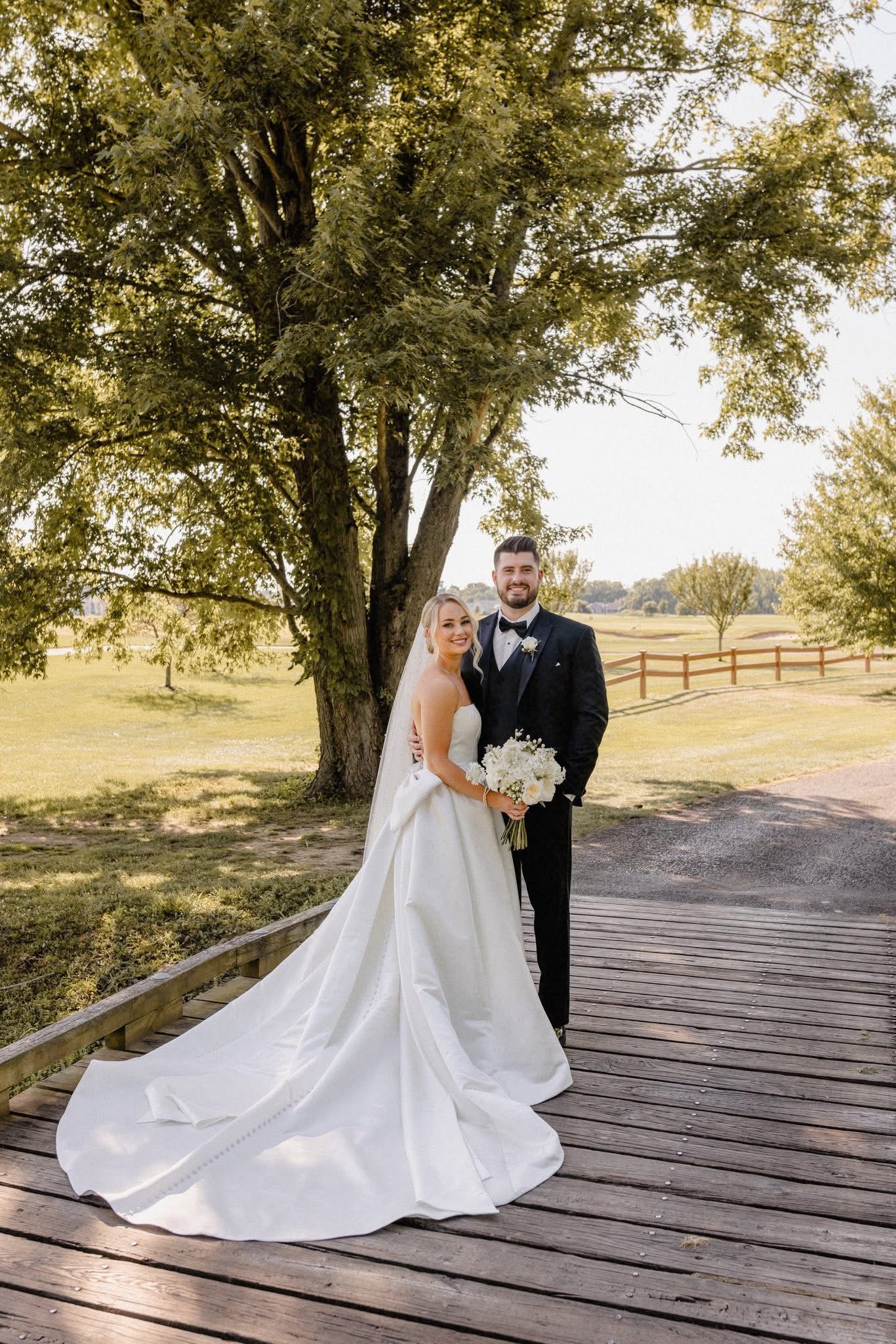 Bride and groom on a wooden bridge, holding flowers. Groom wears a tuxedo. Bride wears a white dress. Green trees and field in background.