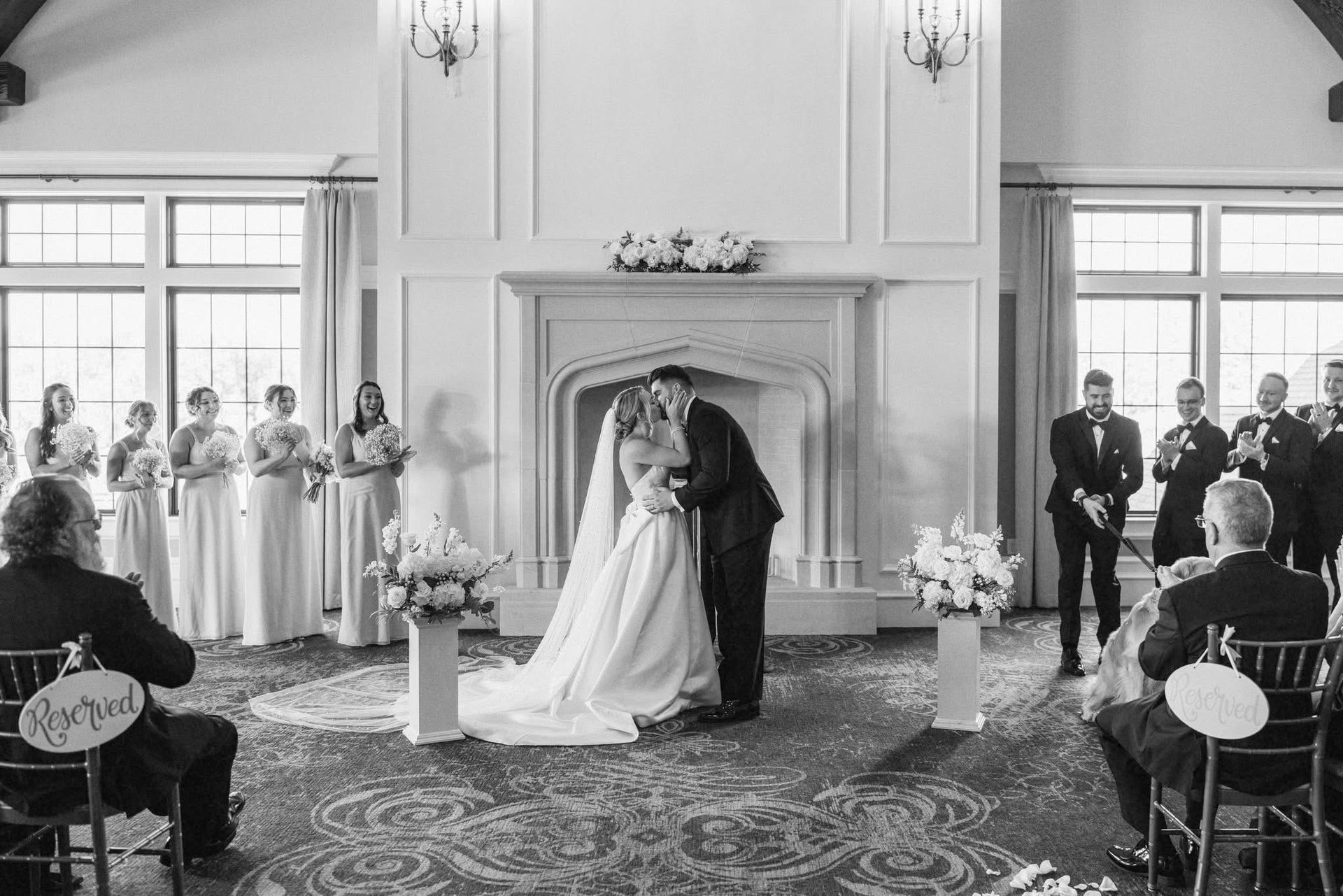 Bride and groom kiss at altar during wedding ceremony, surrounded by wedding party and guests.