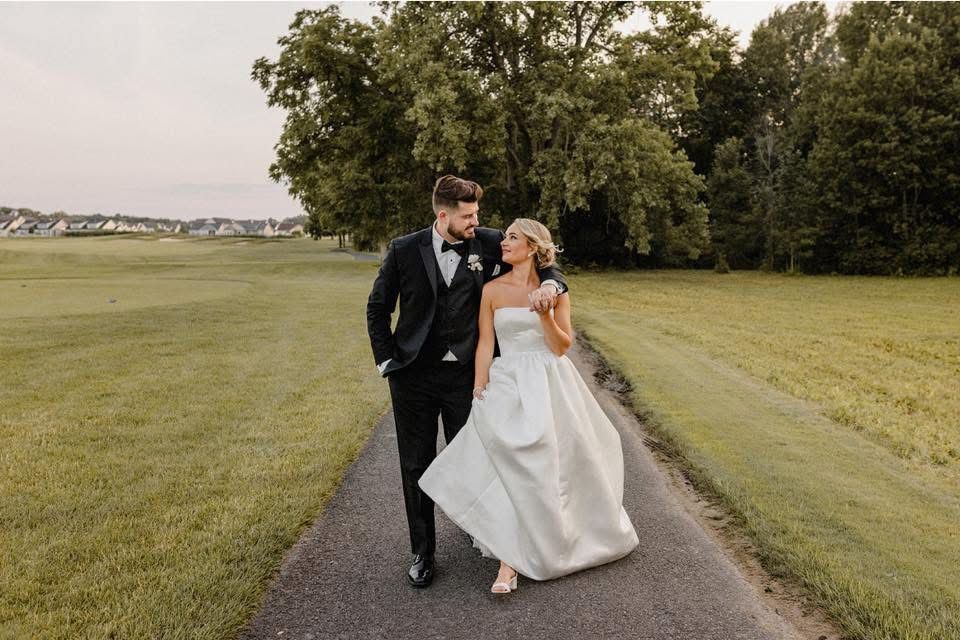 Bride and groom walk down a path, arm in arm. She wears a white gown, he wears a tuxedo. Green fields surround them.