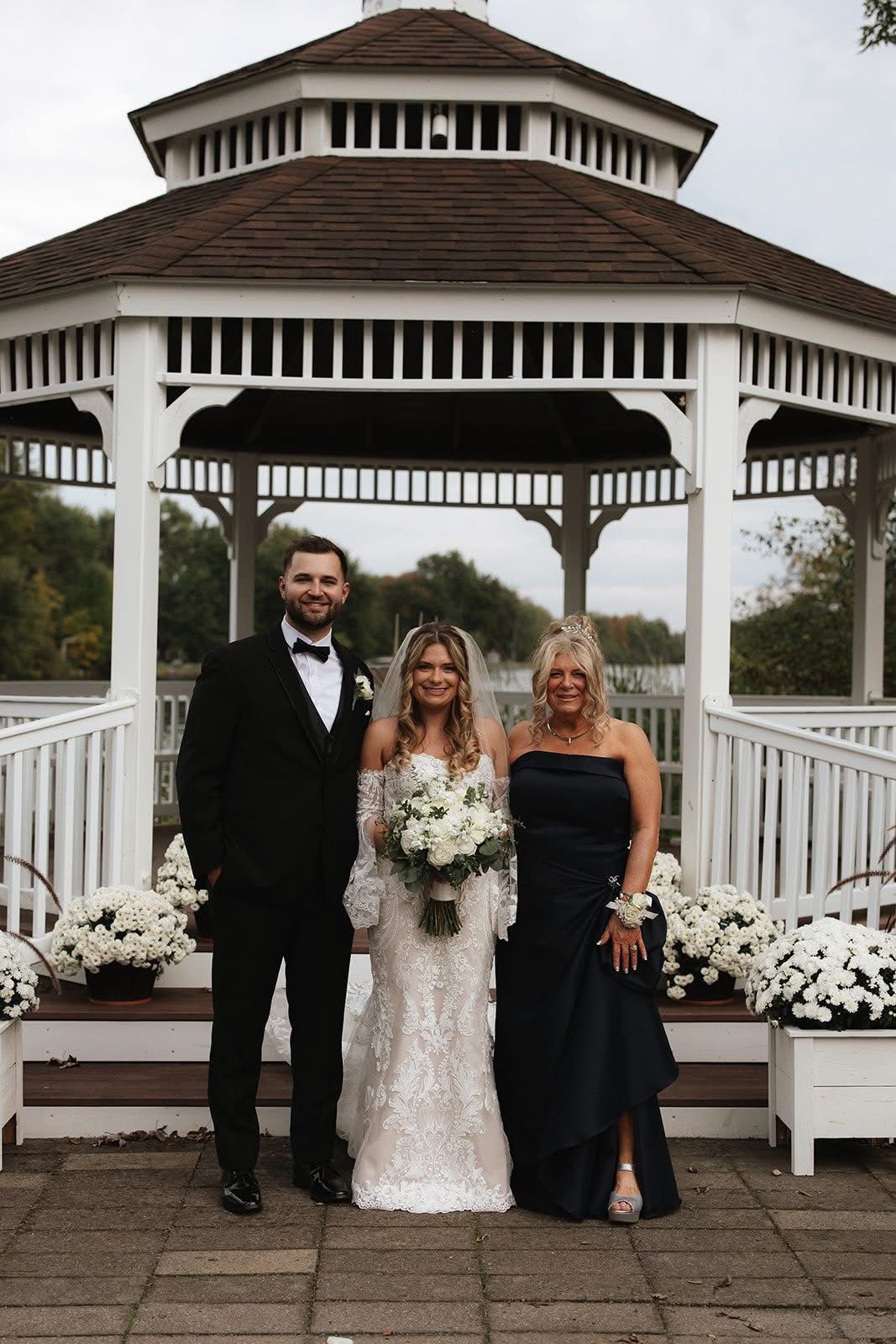 Wedding portrait: Couple and woman in front of a gazebo with white flowers. Man in black suit, woman in white dress, other woman in blue dress.