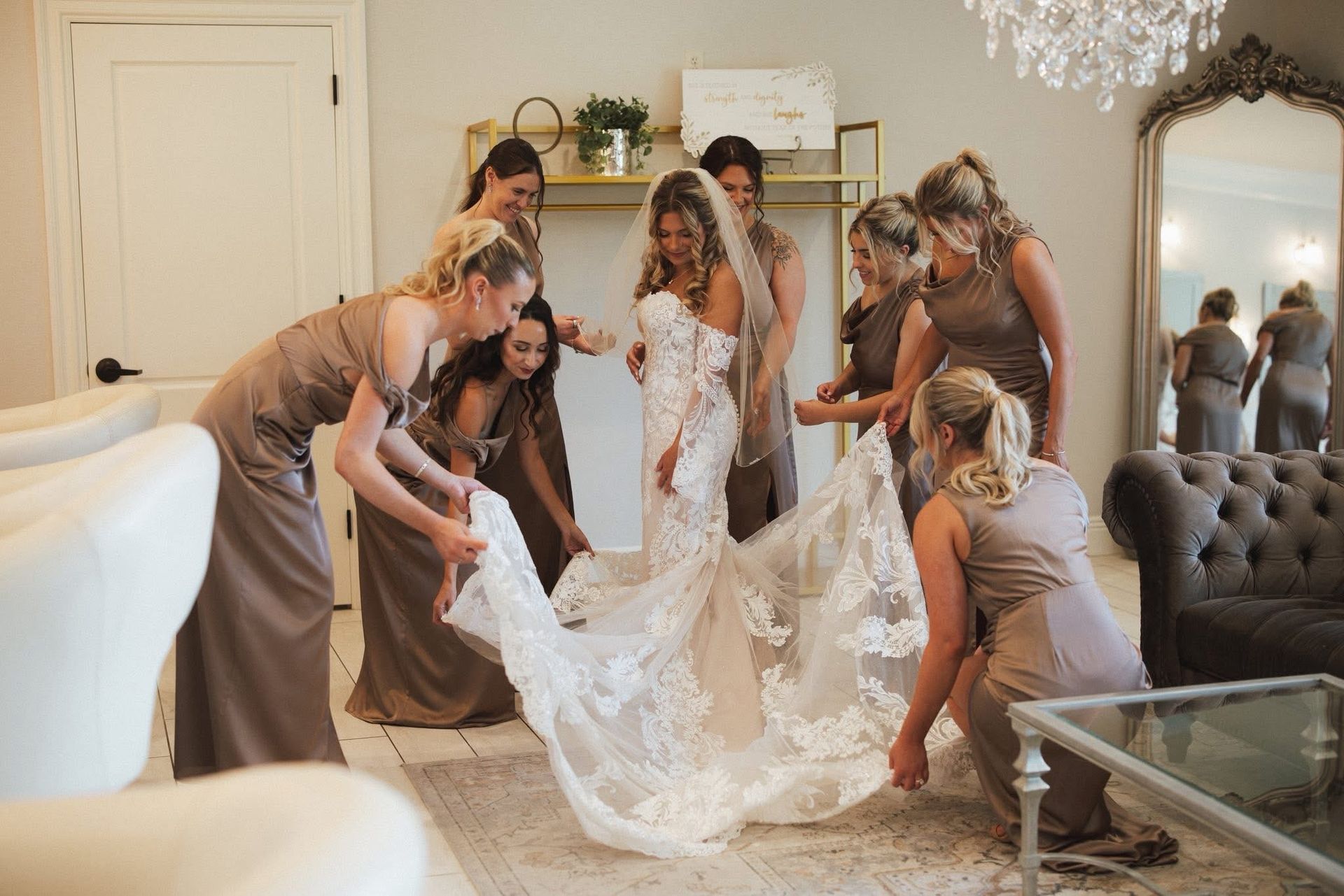 Bride surrounded by bridesmaids, adjusting her long train in a room with a large mirror.