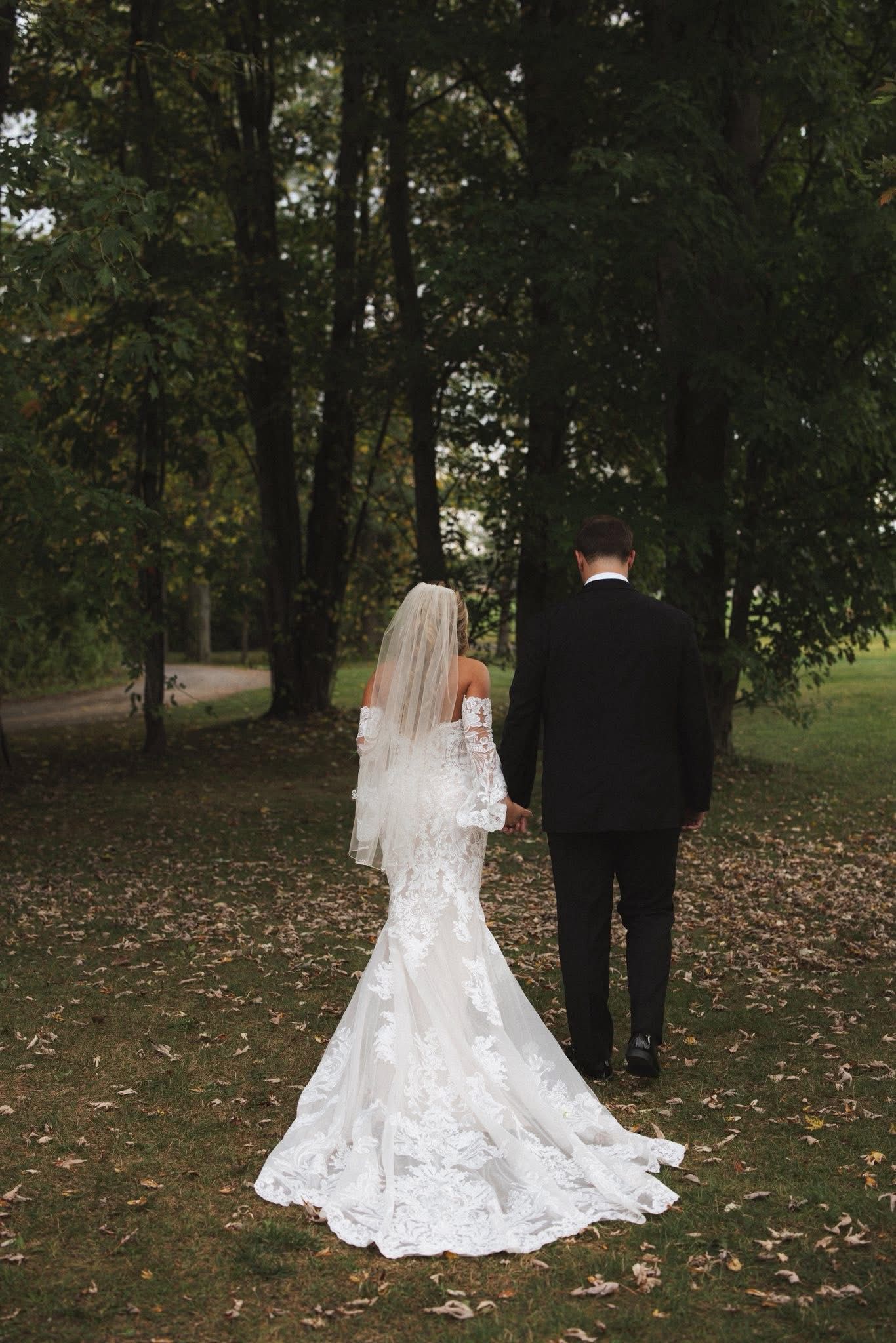 Bride and groom hold hands walking in a park, trees in the background. Bride in white lace dress and veil.