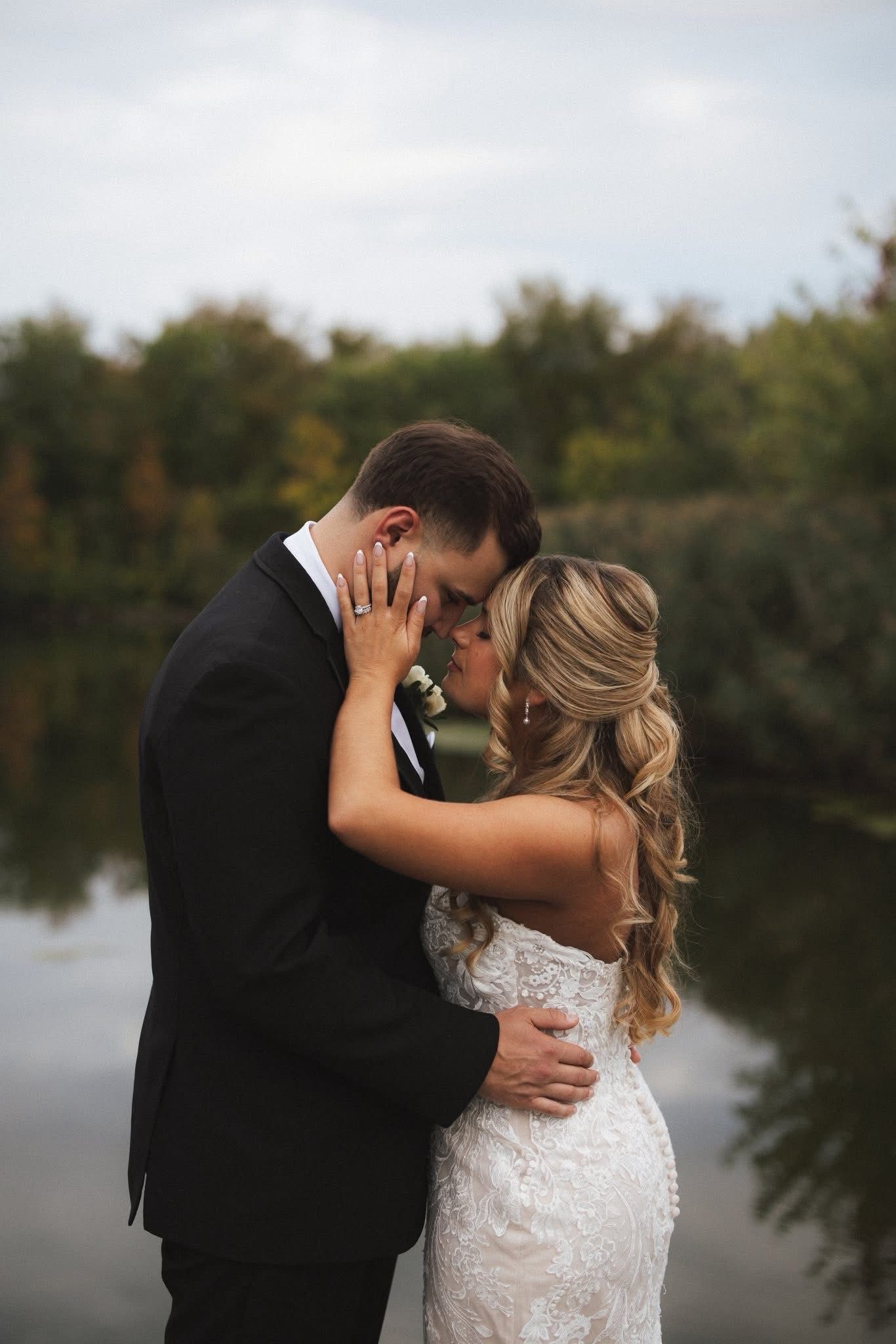 Couple embraces outdoors near water, man in black suit, woman in white dress.