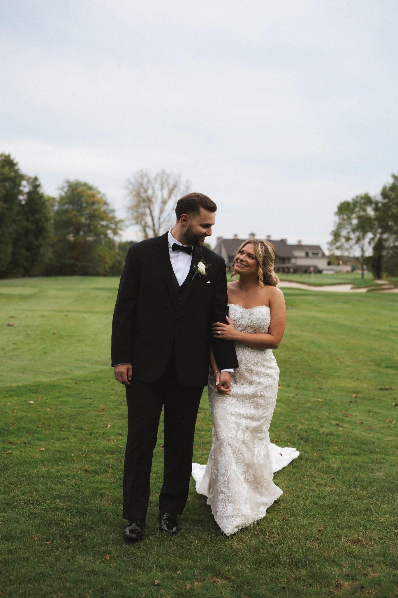 Bride and groom walking on golf course, holding hands, smiling at each other. The groom wears a black suit, and the bride wears a white gown.