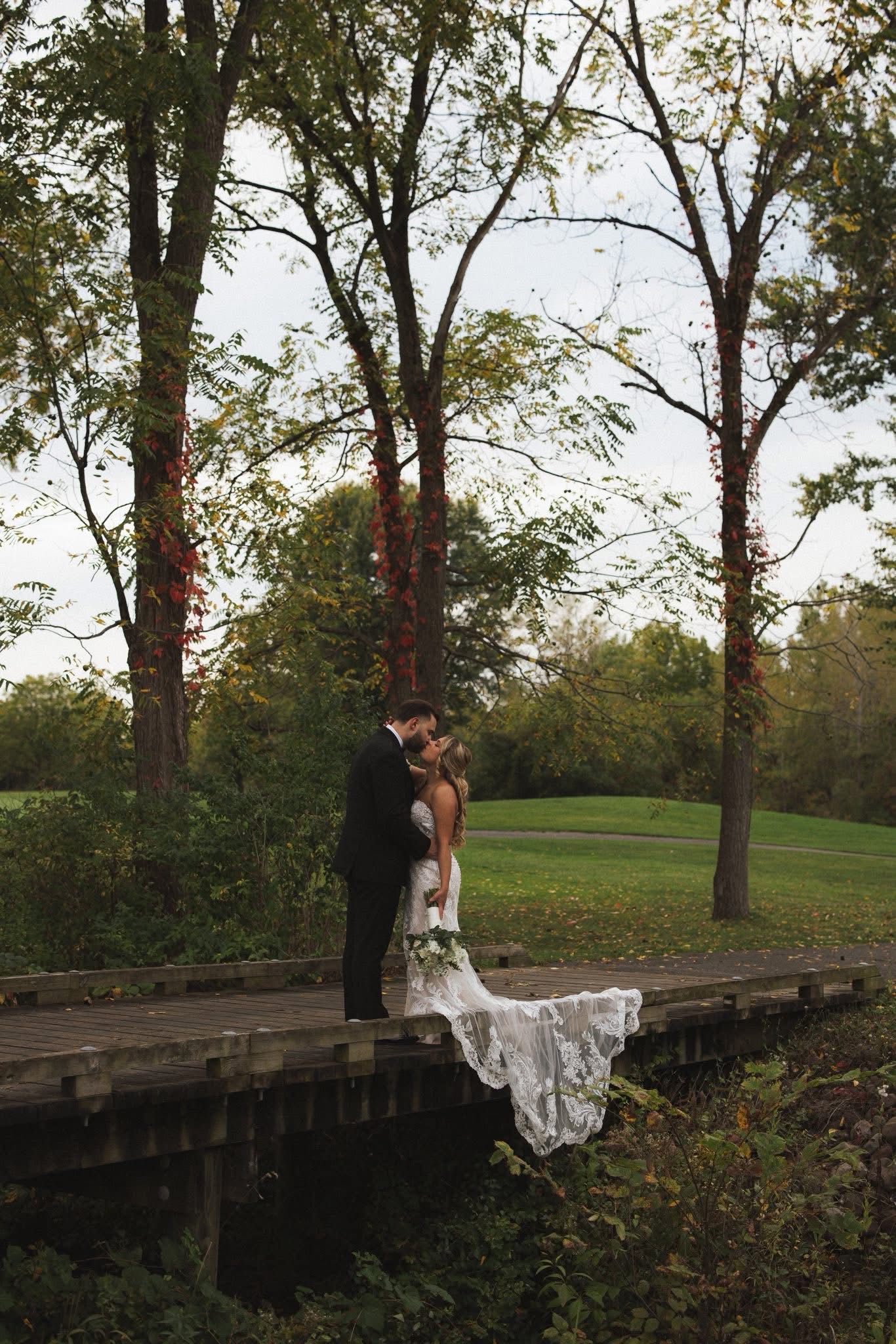 Couple kissing on a wooden bridge, wearing wedding attire. Surrounded by trees and greenery.
