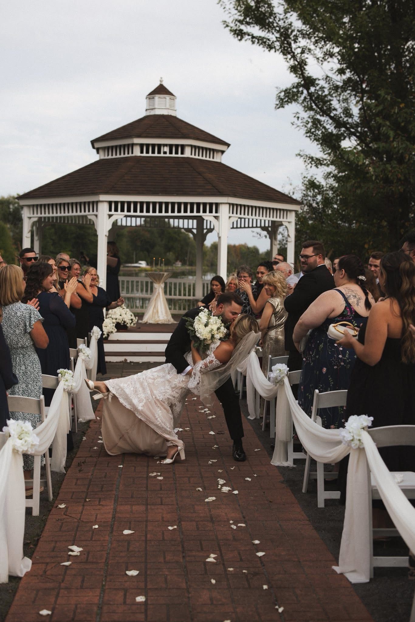 Bride and groom embrace on brick aisle after ceremony, guests cheer near gazebo and lake.
