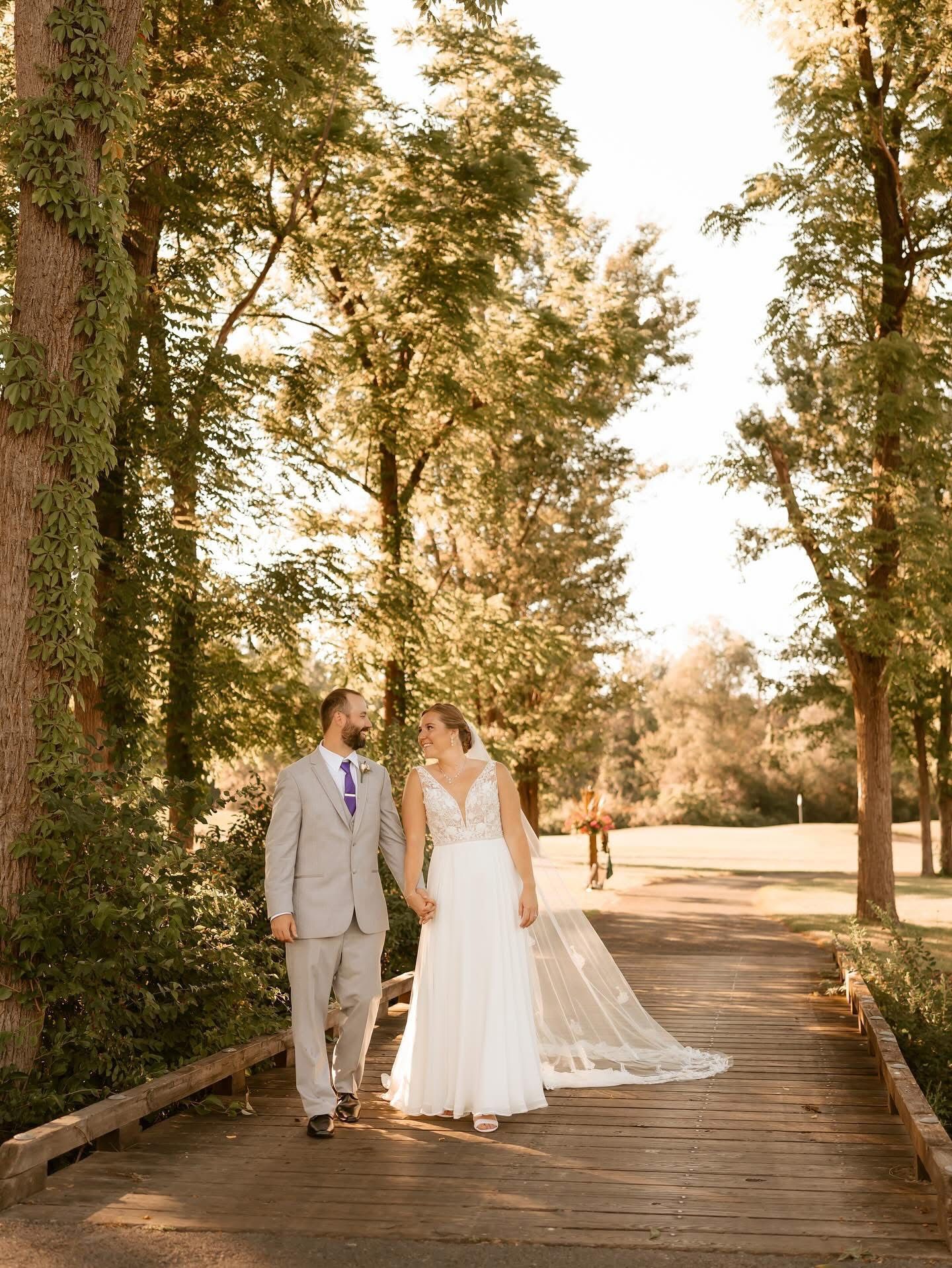 Bride and groom hold hands, walking on a wooden bridge in a sunlit outdoor setting.