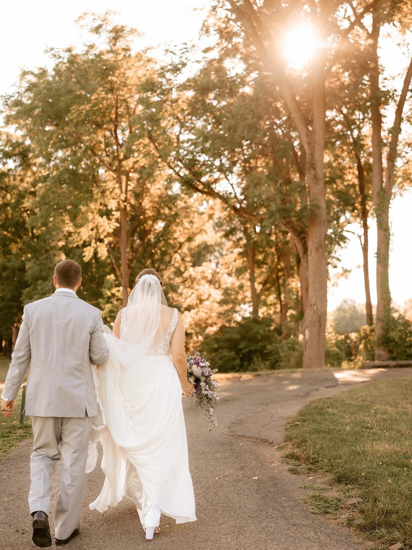 Bride and groom walk away, holding hands, down a path. Sunlight shines through trees.