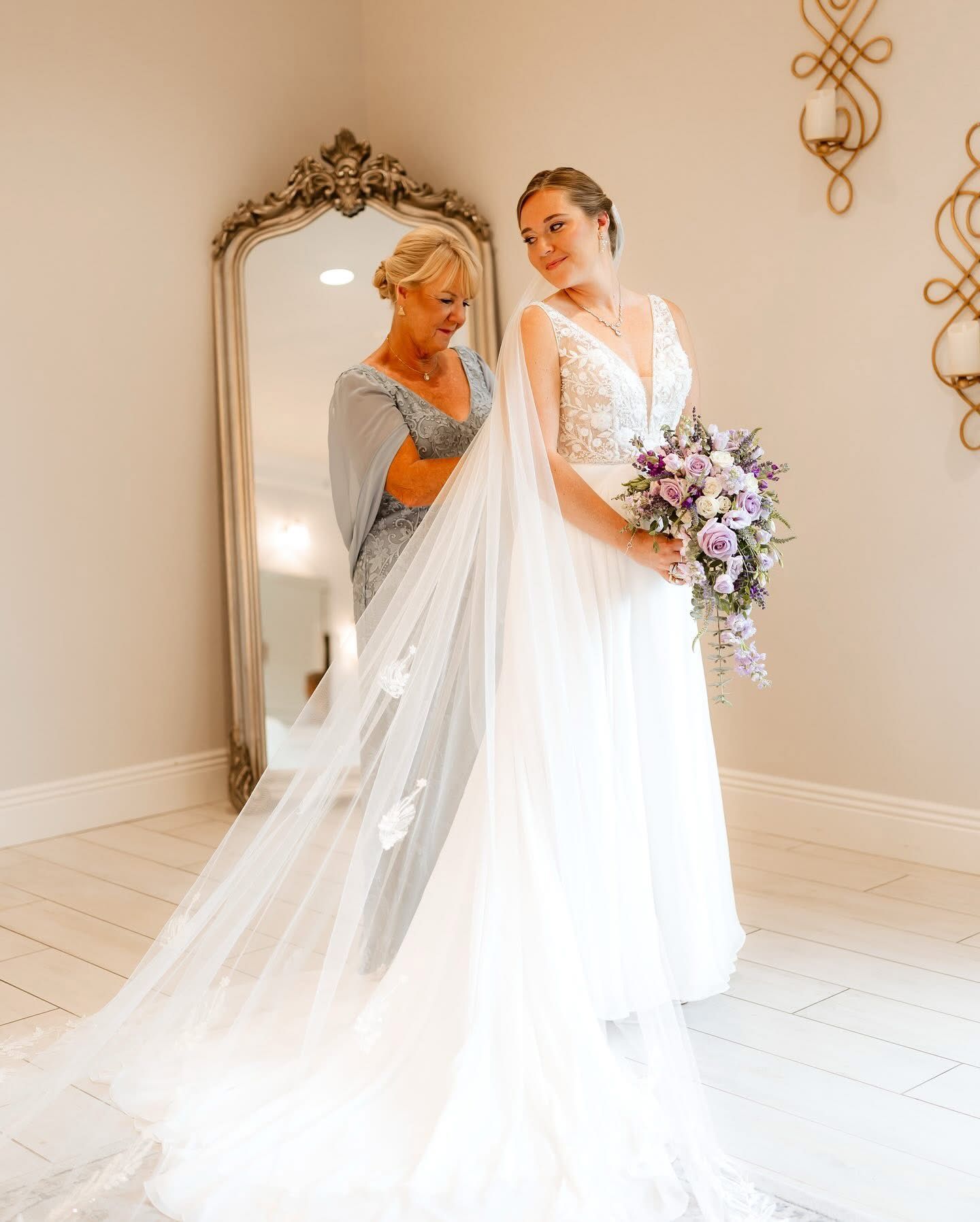Bride in a white dress with a veil, standing with her mother who adjusts her veil in a room with a large mirror.