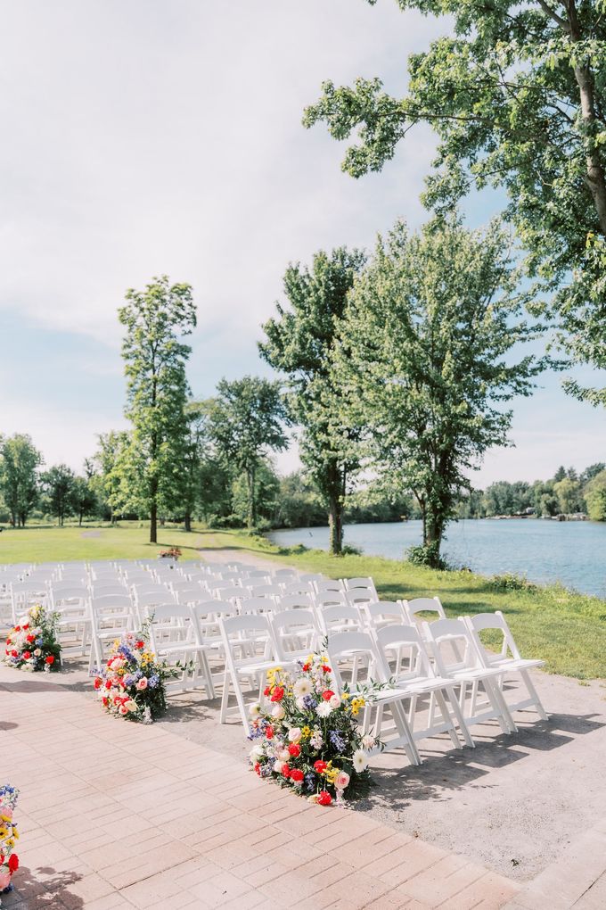 White chairs arranged for a lakeside ceremony, decorated with colorful flowers; sunny day.