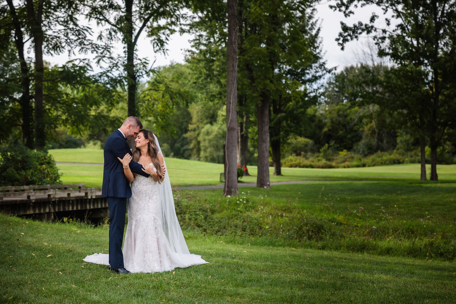 Couple embracing on a grassy lawn with a small wooden bridge and trees in the background. The man kisses the woman's head.