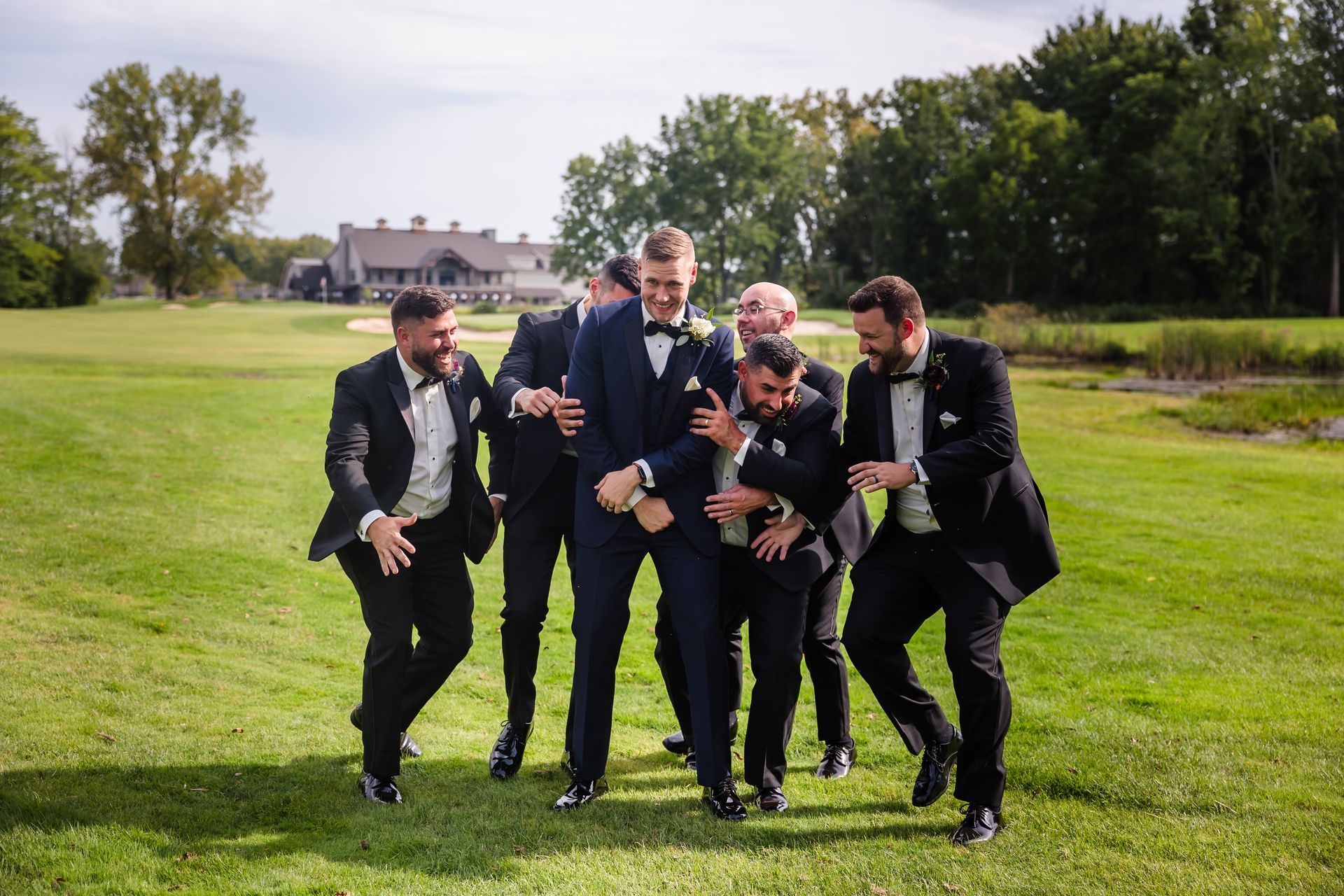 Men in tuxedos playfully surround a man in a navy suit on a green golf course, smiling.