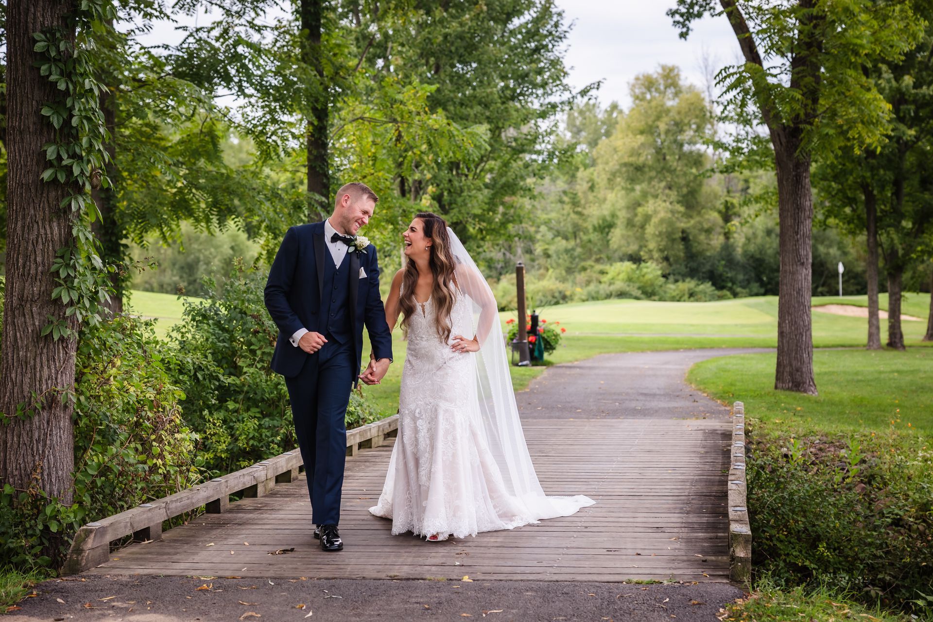Bride and groom walking and laughing on a wooden bridge, surrounded by greenery.