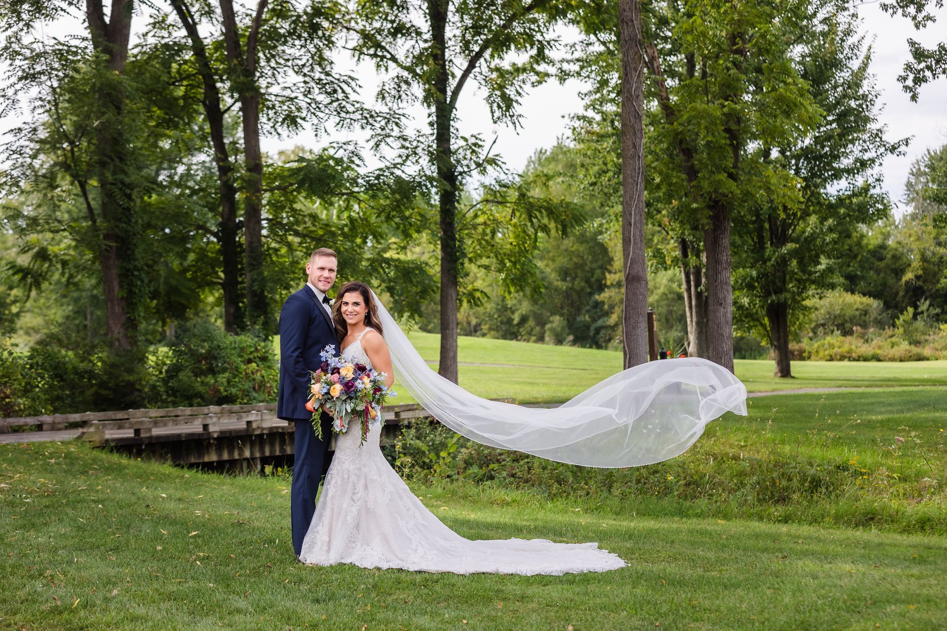 Bride and groom in formal attire pose on a grassy lawn; bride's veil flowing.
