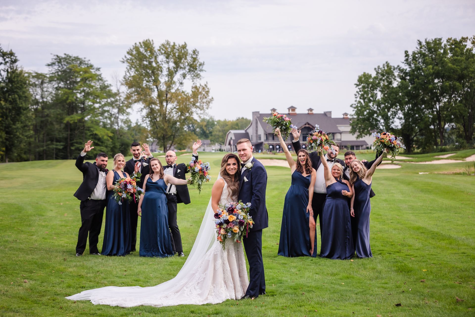 Wedding party on a golf course. Bride and groom in front; others wave, holding bouquets.