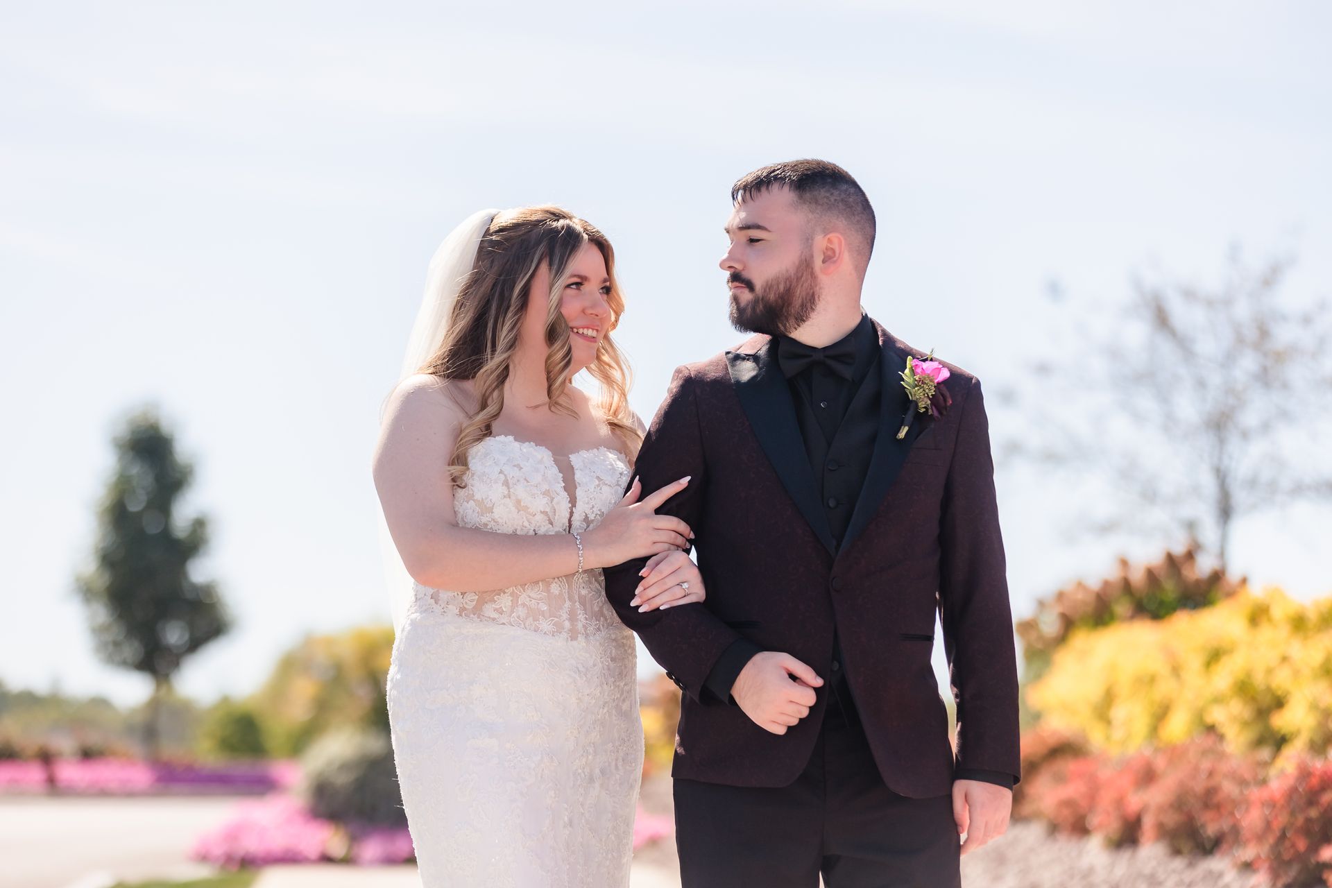 Newlyweds walk arm-in-arm outside, looking at each other. The bride wears a white gown, and the groom a dark suit. Bright sunny day.