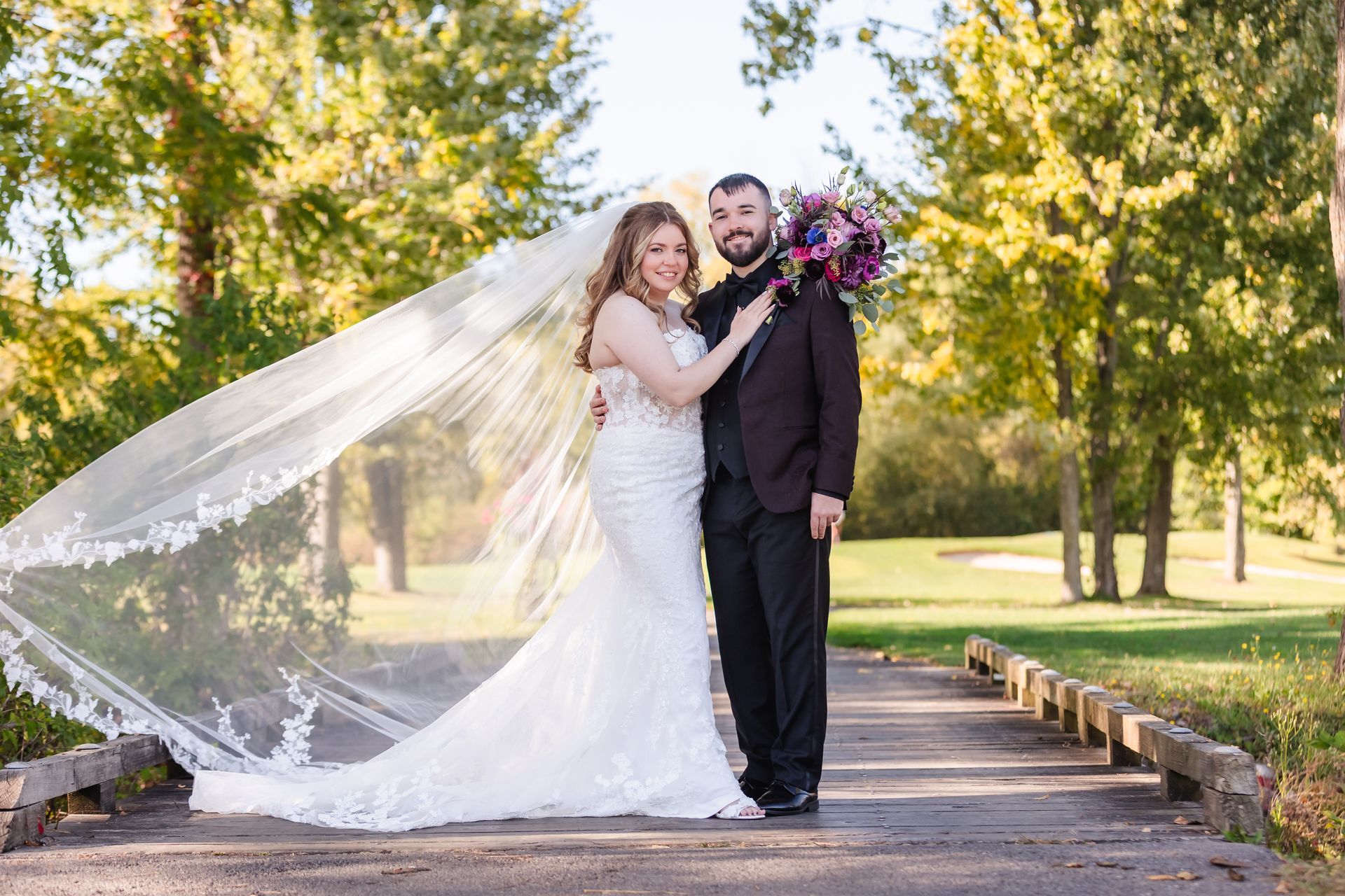 Bride and groom stand on a bridge, with the bride's veil flowing. Autumn trees in the background.