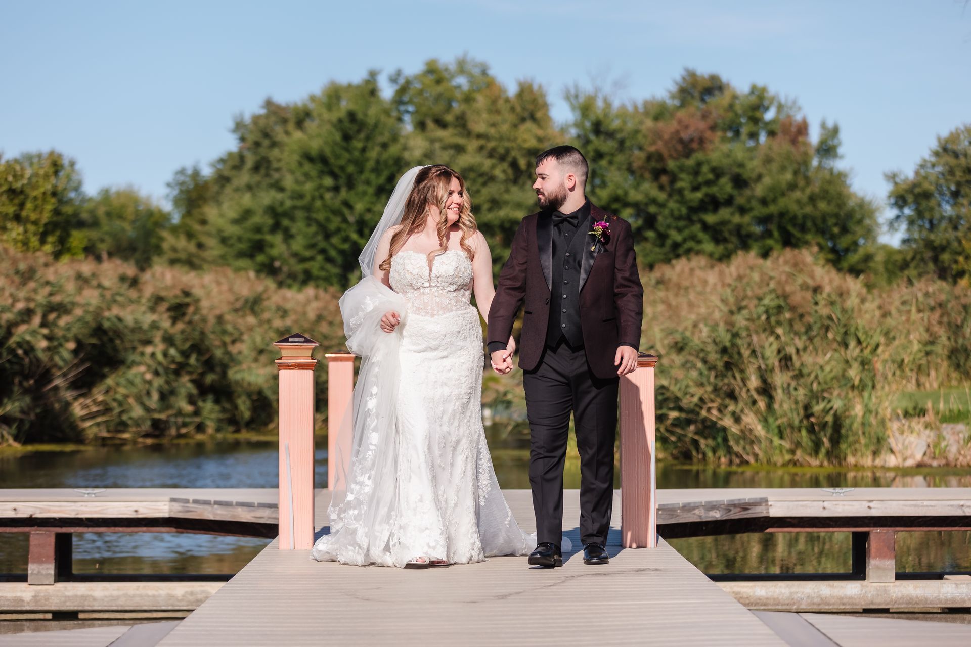 Bride and groom holding hands, walking on a pier, looking at each other, trees and water in the background.