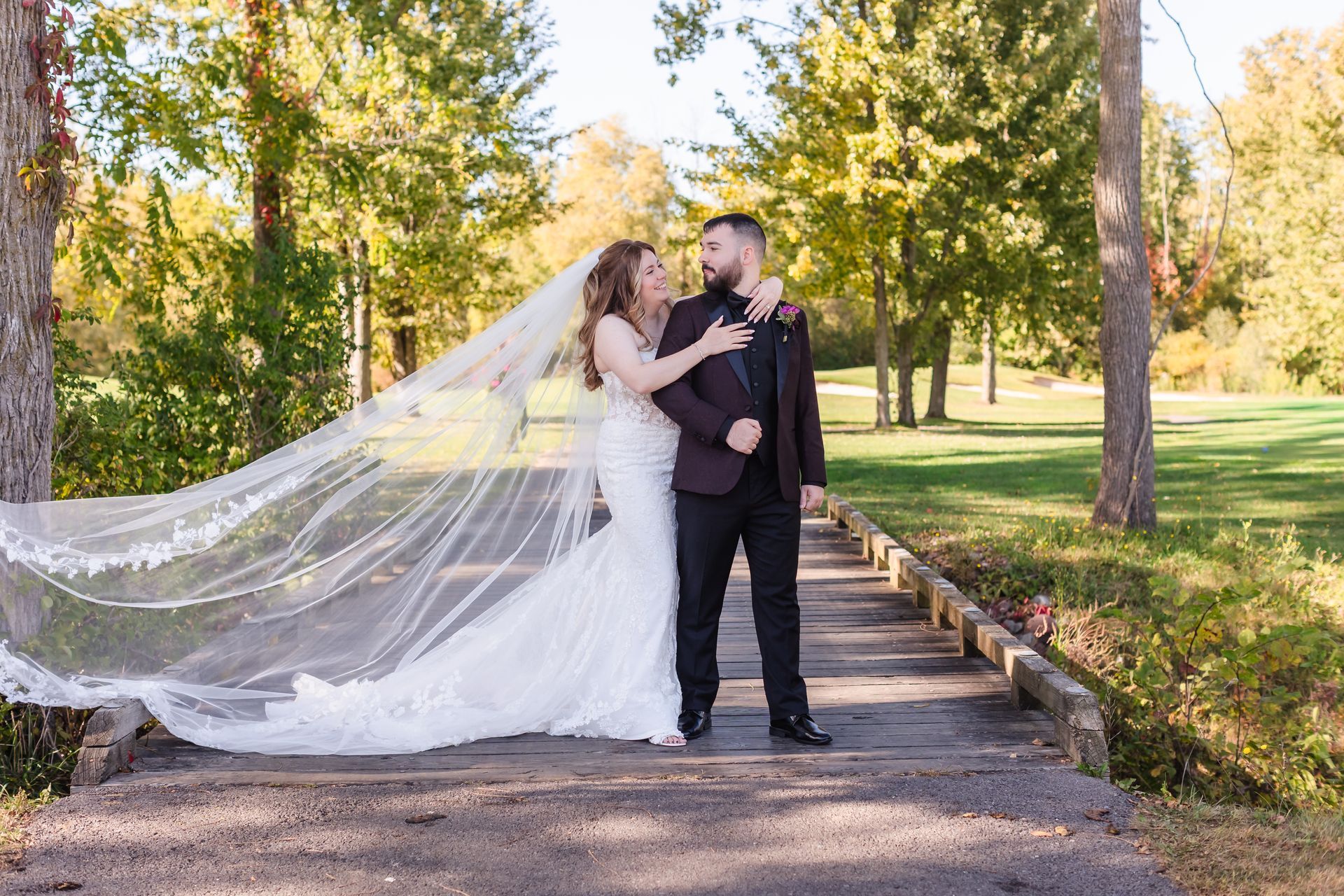 Bride and groom pose on a bridge outdoors, bride's veil flowing. Autumn trees in the background.