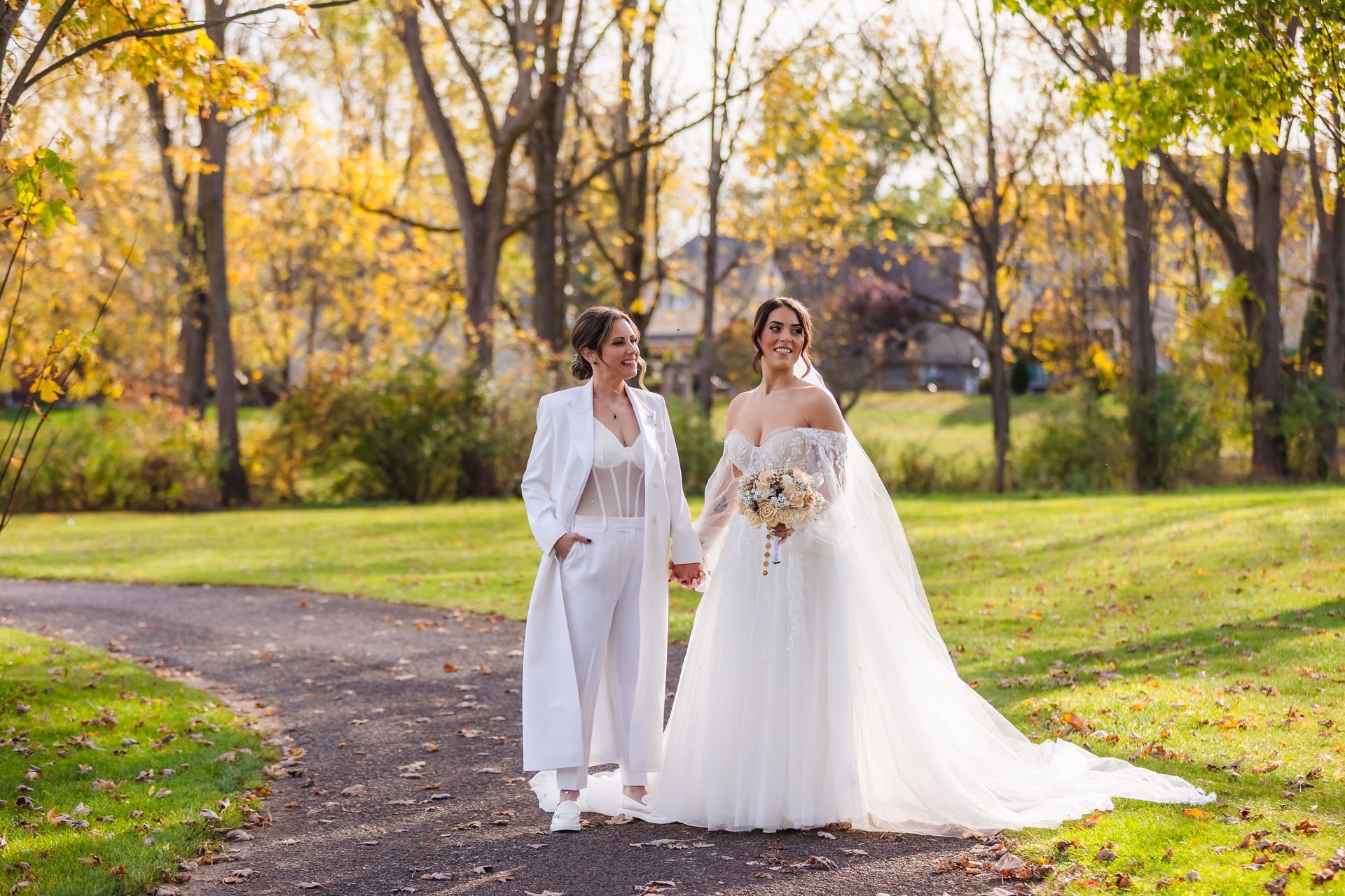 Two brides holding hands, walking on a path in a park; one in a white pantsuit, the other in a white gown.