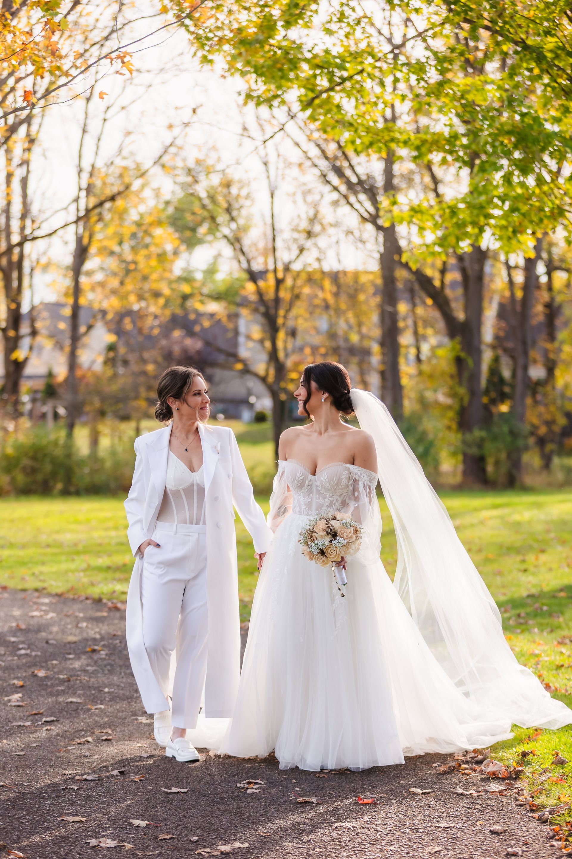 Two women, one in a white suit, the other in a wedding dress, hold hands and walk on a path, outdoors.