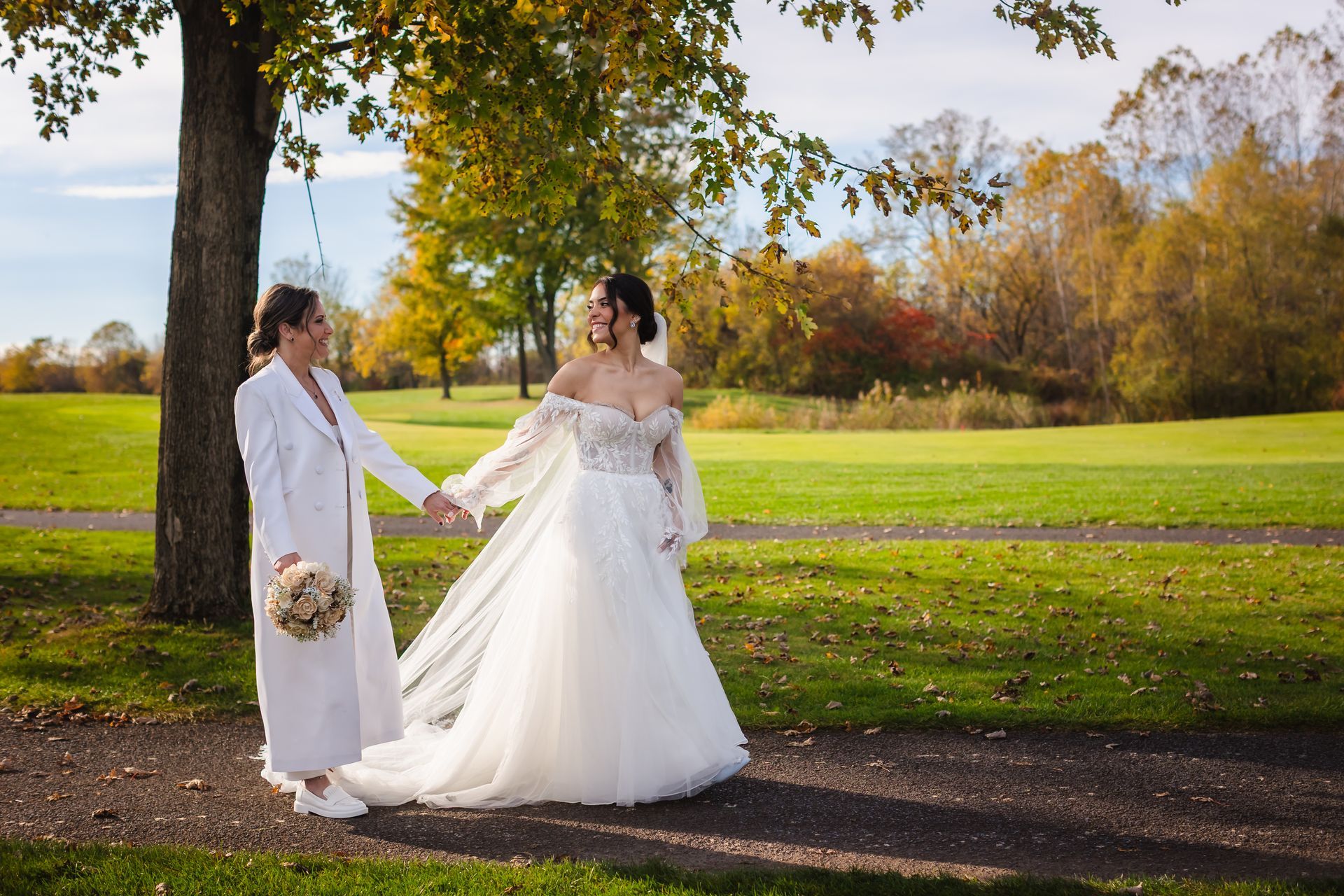 Two people, likely a couple, holding hands in a park. One in a white dress, the other in a white suit, smiling.