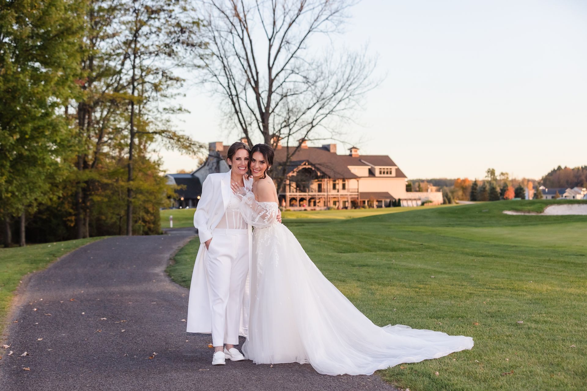 Two women in wedding attire pose on a paved path, a large house and golf course in the background.