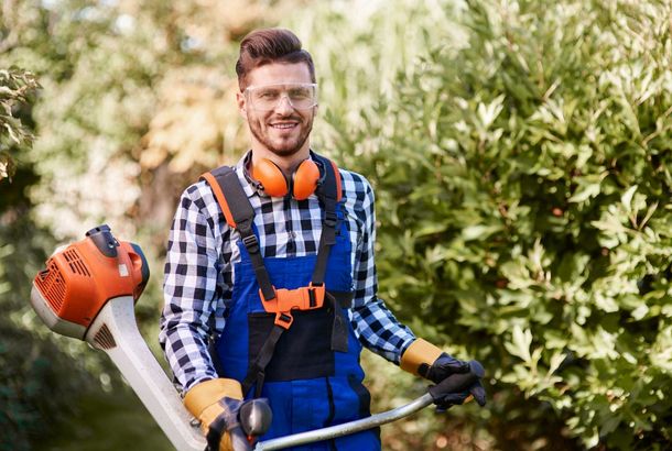 Person in blue overalls holding a string trimmer in a sunny garden, with orange safety gear.