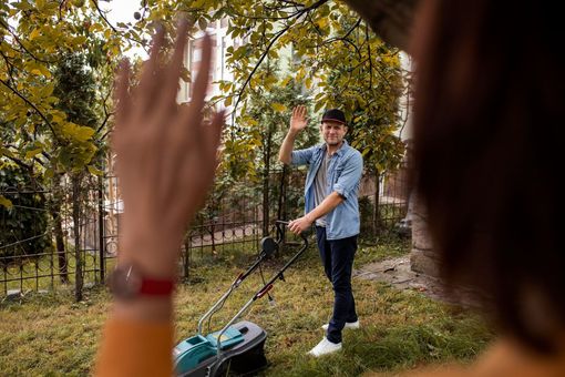 Two people waving in a wooded park, one in the foreground and one near a lawn mower.