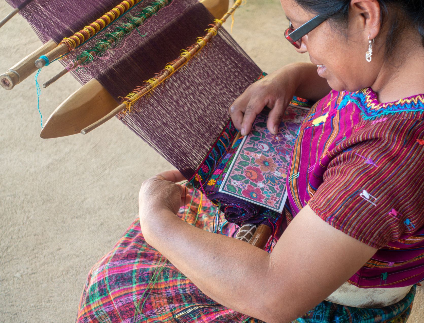 Woman weaving colorful textile on a loom; she's wearing traditional clothing, indoors.