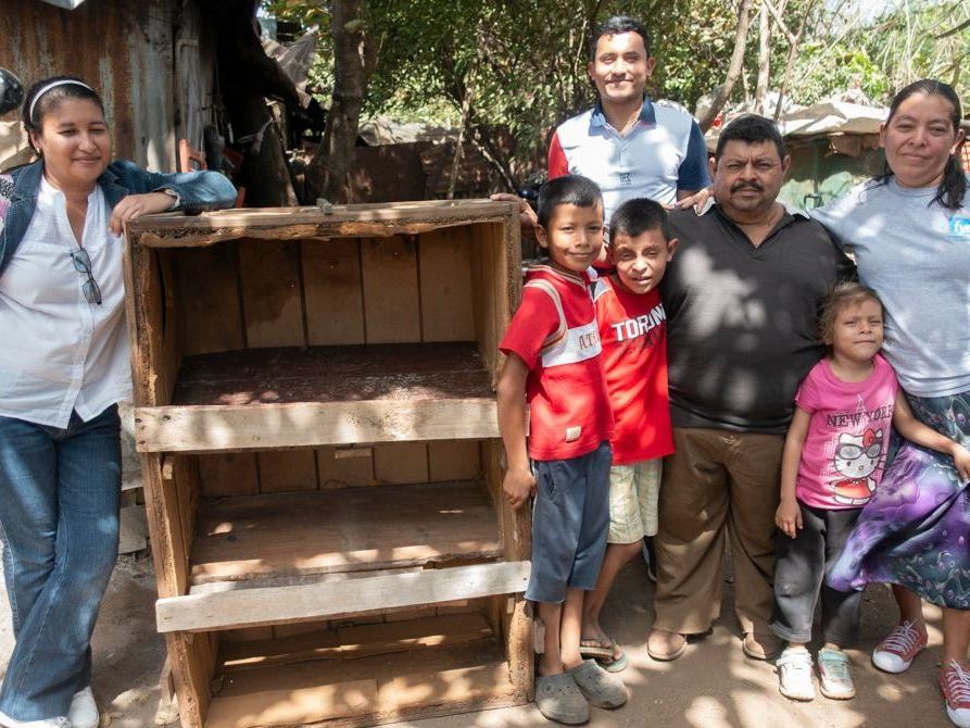 Family stands by a wooden composting bin; children in red, adults smiling in outdoor setting.