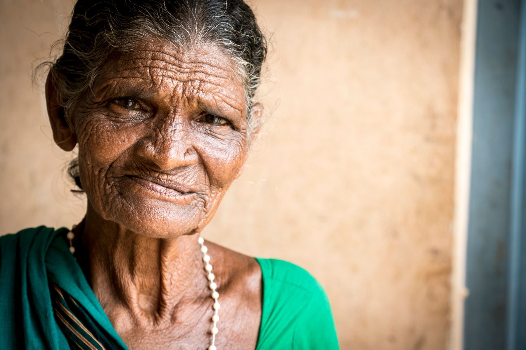 Elderly woman with wrinkled skin and a slight smile, wearing a green top and necklace against a light-colored wall.