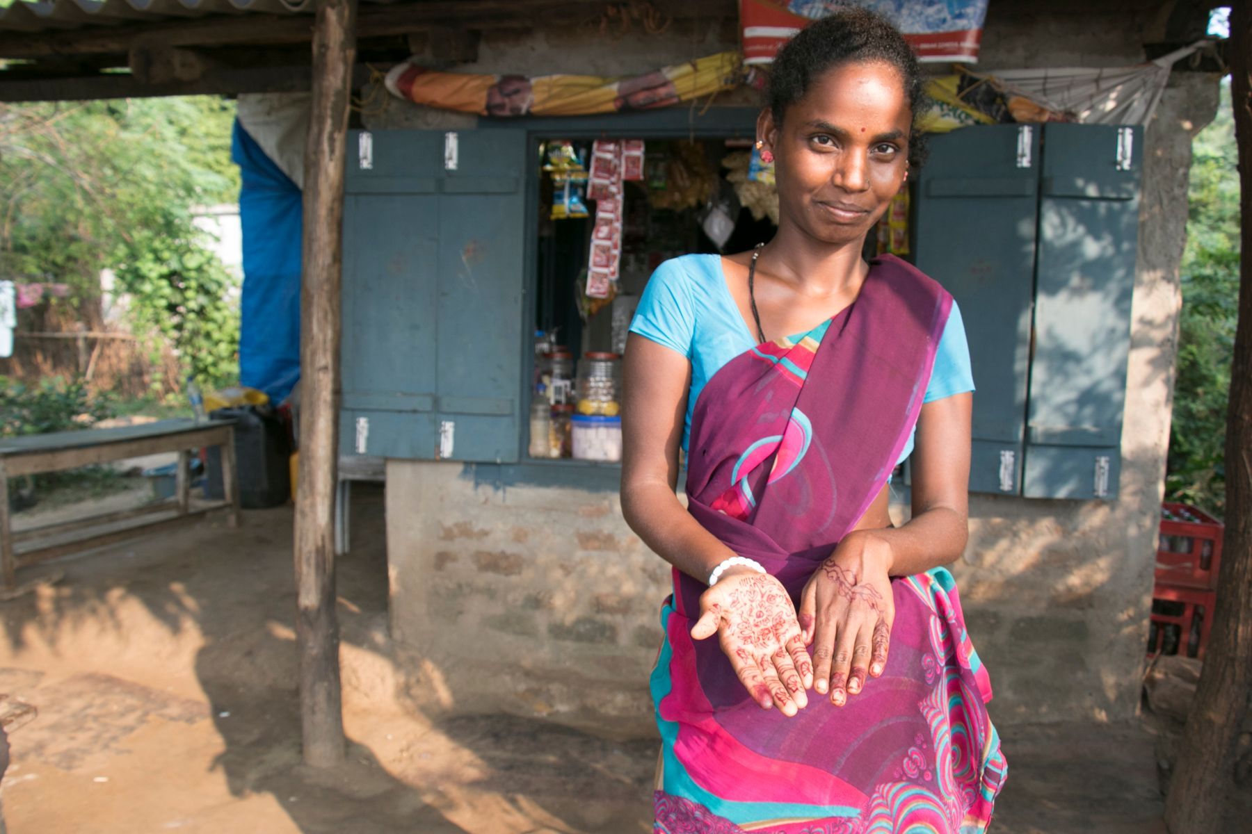 Woman in a sari stands in front of a small shop with her palms facing forward, smiling.