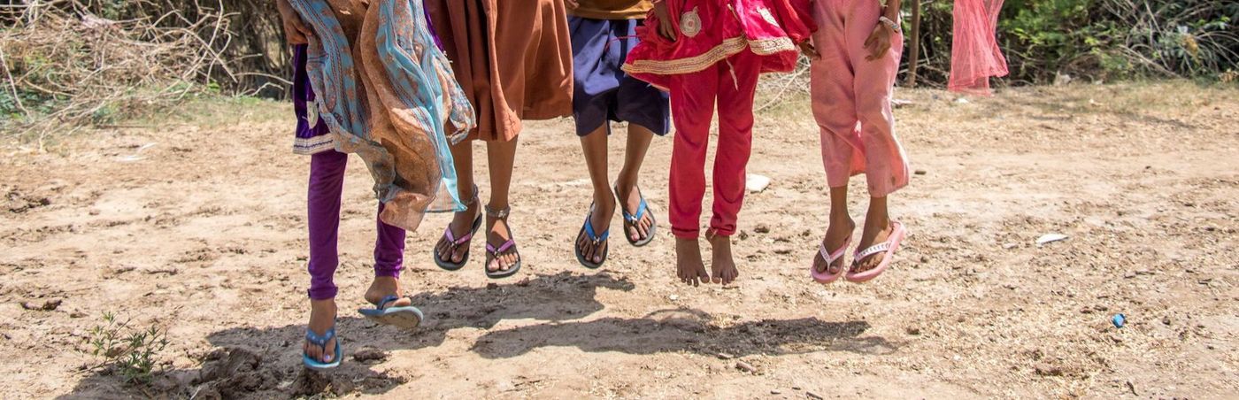 Children jumping in the air, legs in colorful clothing, on a sandy surface.