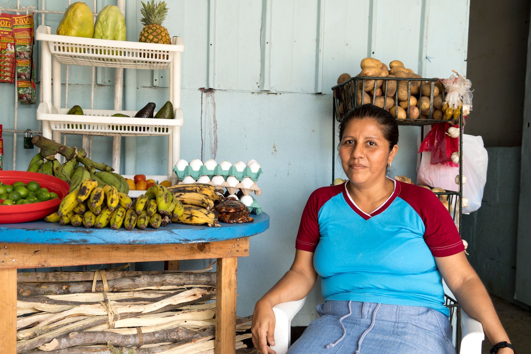 Woman at a fruit stand, blue shirt, various fruits and vegetables displayed on a wooden table, light blue wall.