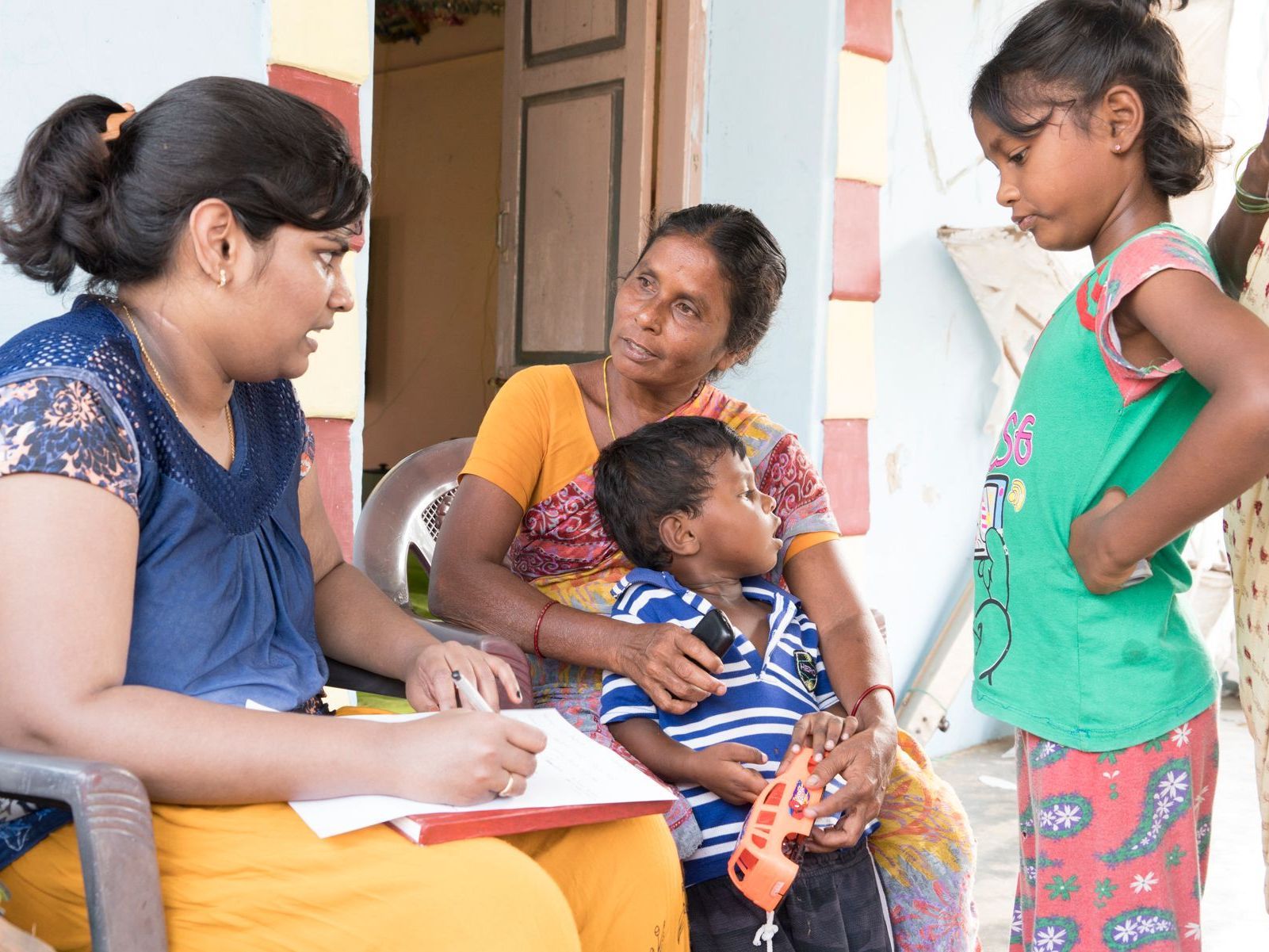 Woman interviewing a family: Mother, child, and older girl. Outdoors, bright colors.