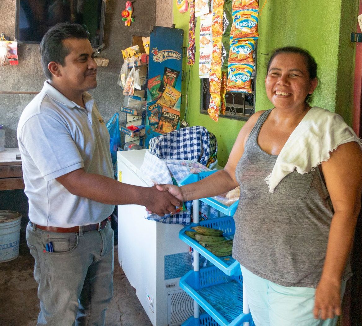 Man and woman shake hands in a store; woman smiles, shelves, products, refrigerator in background.