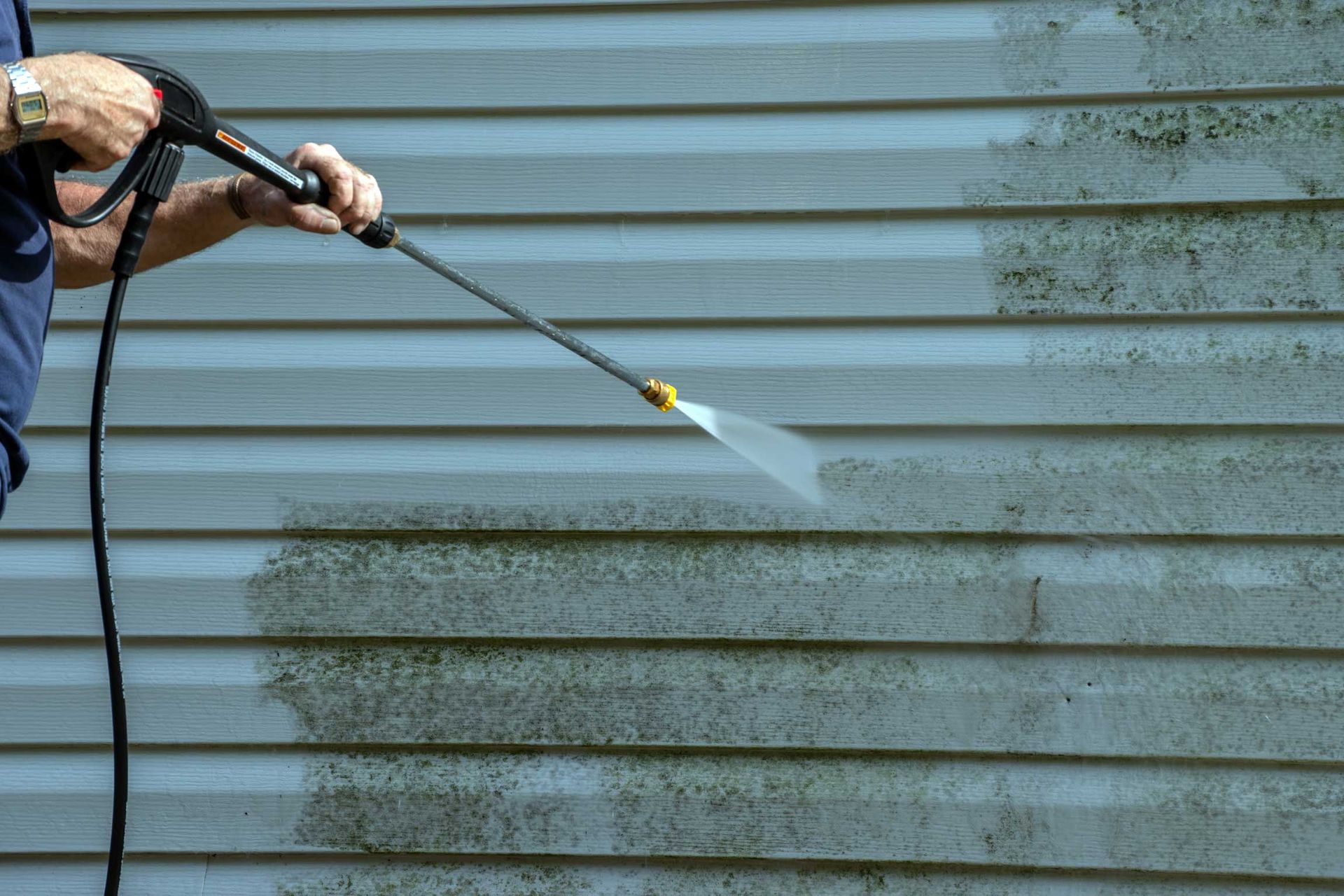 Person pressure-washing a dirty white siding wall with a spray wand
