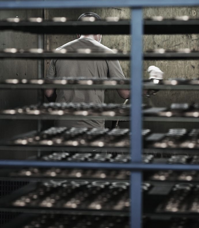 Person working in a factory setting behind metal racks, shelves filled with trays. The person is wearing light colored clothing and is holding something in their right hand.
