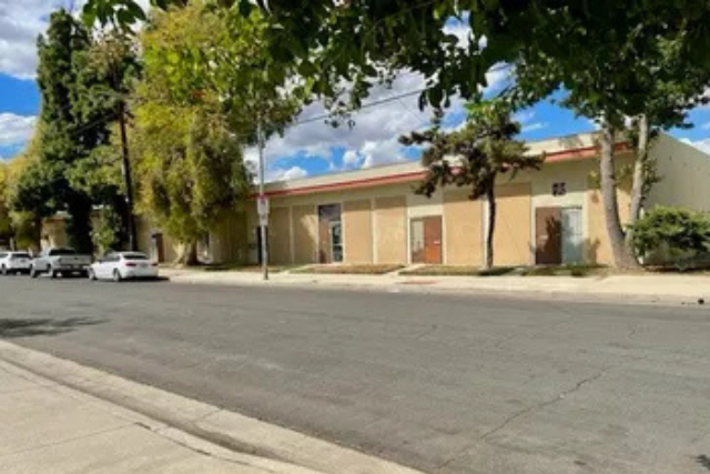 A wide view of a quiet street lined with tall trees and a beige commercial building under a bright sky with scattered clouds.