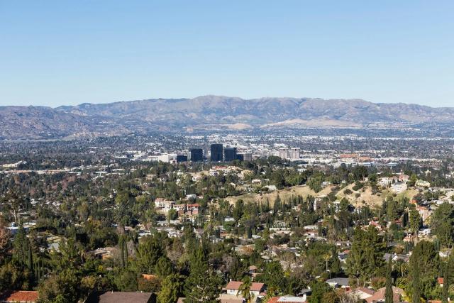 A wide view of a city surrounded by trees, with tall mountains in the background under a clear blue sky.