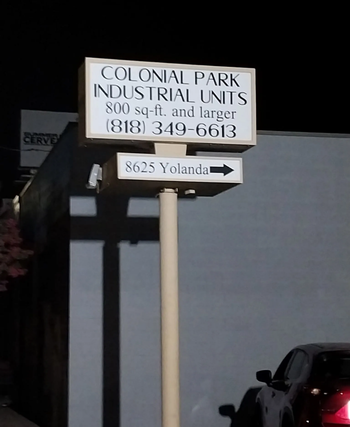 A tall sign at night showing the text “Colonial Park Industrial Units, 800 sq-ft and larger, (818) 349-6613” with a smaller sign below it pointing toward “8625 Yolanda.” A dark building and a parked car are in the background.