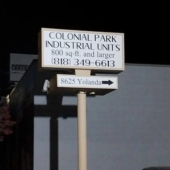 A tall sign at night showing the text “Colonial Park Industrial Units, 800 sq-ft and larger, (818) 349-6613” with a smaller sign below it pointing toward “8625 Yolanda.” A dark building and a parked car are in the background.